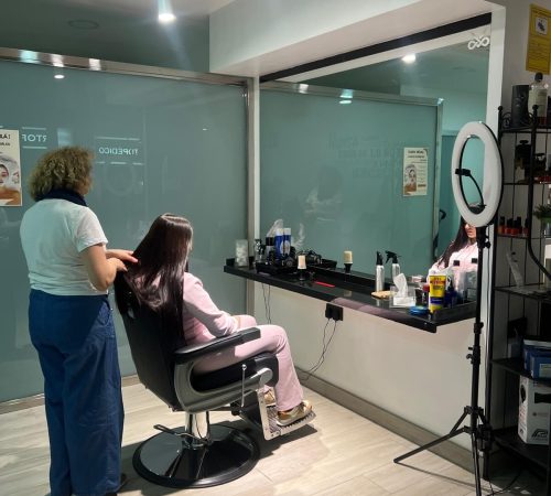 Estilista realizando un tratamiento capilar a una clienta en un salón moderno, con productos de belleza visibles en la mesa y un espejo reflejando el ambiente.